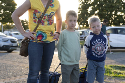 une femme et deux enfants debout sur le trottoir, les deux garçons tiennent leur cartable comme une valise dans leur dos - Agrandir l'image 72 sur 80, fenêtre modale