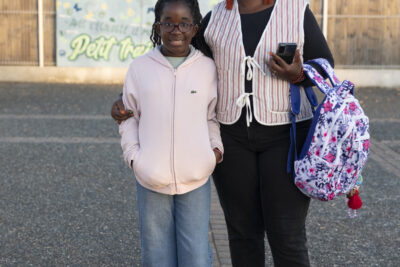 une femme et sa fille debout et souriantes, la mère tient le sac à dos de sa fille au bras, elle tient le bras de sa fille de l'autre main, derrières elles on voit la fresque bleue avec écrit "Petit train", le nom de l'école - Agrandir l'image 66 sur 80, fenêtre modale