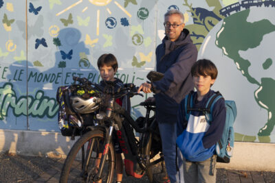 un père et ses deux fils, il est à côté de son vélo et un enfant de chaque côté, devant la fresque bleue de l'école - Agrandir l'image 69 sur 80, fenêtre modale