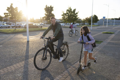 un homme à vélo et une jeune fille à trottinette, sourient, à l'arrière plan on voit une jeune fille de dos à vélo, et le parking et le soleil levant - Agrandir l'image 71 sur 80, fenêtre modale