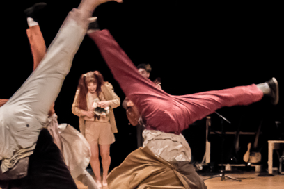 plan rapporché sur les circassiens du spectacle en train d'effectuer des grands écrats tête renversés, en arrière-plan, debout la silhouette d'une comédienne tête baissée en train de pouffer de rire, vêtue d'une toge et d'une couronne dans les tons de beige - Agrandir l'image, fenêtre modale