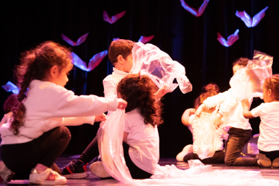 photo d'enfants en chemise blanche et pantalon noir assis de part et d'autre d'une scène de spectacle, sous les lumières rosées et l'habillage lumineux figurant des oiseaux en vol - Agrandir l'image, fenêtre modale