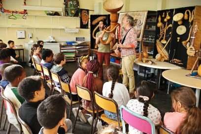 vue d'ensemble d'engfants assis sur leur chaise de classe face à deux musiciens sur une estrade en tenues décontractées jouant de leur instrument à vent - Agrandir l'image, fenêtre modale