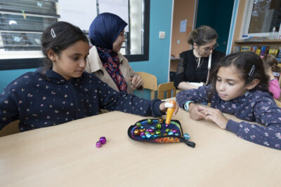 photo de jeunes filles qui jouent à un jeu sur une table, on voit deux femmes assises derrière également - Agrandir l'image 10 sur 19, fenêtre modale