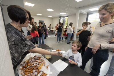 photo paysage de la pièce où a lieu la fête avec au premier plan une femme qui donne un beignet à un petit garçon accompagné d'une femme, on voit des familles à l'arrière plan en train de discuter ou de jouer à des jeux - Agrandir l'image 6 sur 19, fenêtre modale