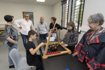 photo d'un groupe de personnes qui discutent dans une pièce autour de deux enfants qui jouent à un jeu en bois posé sur une table - Agrandir l'image 19 sur 19, fenêtre modale