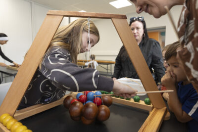 photo paysage d'une jeune fille qui joue à un jeu de bois, une structure posée sur une table avec des boules qui tiennent ensemble au centre en équilibre, d'autres enfants et personnes regardent autour - Agrandir l'image 13 sur 19, fenêtre modale