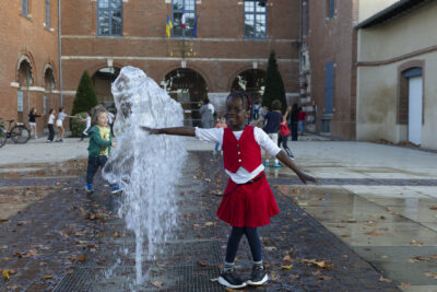 photo d'une petite fille habillée en rouge et blanc qui joue avec les jets d'eau sur le parvis de la Mairie et sourit à l'objectif - Agrandir l'image 14 sur 20, fenêtre modale