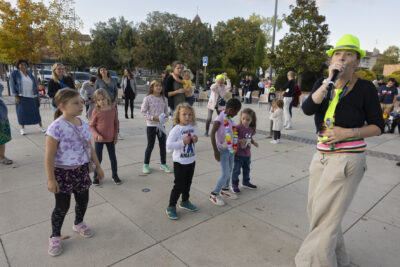 photo d'enfants qui dansent sur le parvis de la Mairie, guidés par une femme qui tient un micro et porte un chapeau jaune fluo - Agrandir l'image 2 sur 20, fenêtre modale