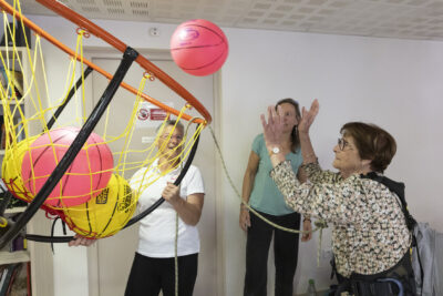 une femme âgée fait du basket avec des ballons légers et un grand panier, devant deux femmes qui l'encouragent - Agrandir l'image 8 sur 20, fenêtre modale