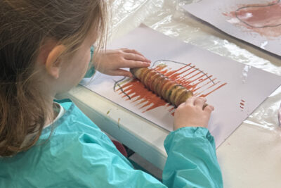 photo d'une jeune fille qui utilise un rouleau à bois avec des sortes de crans pour peindre sur sa feuille blanche avec de la peinture ocre - Agrandir l'image 4 sur 10, fenêtre modale