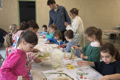 Photo de la table longue avec les enfants assis de chaque côté en train de peindre, ils portent des sortes de tablier pour protéger leurs vêtements et sont concentrés sur leur peinture - Agrandir l'image 7 sur 10, fenêtre modale