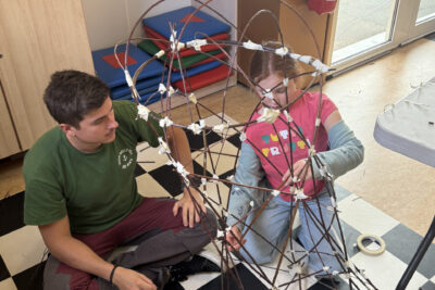 photo d'un animateur et d'une jeune fille par terre en train d'assembler leur lanterne faite de branches fines accrochées ensemble par des scotchs - Agrandir l'image 3 sur 8, fenêtre modale