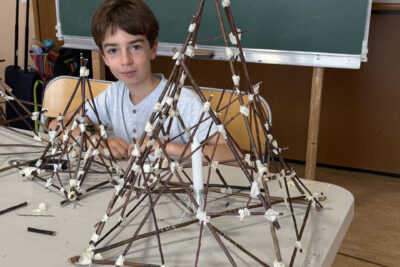 photo d'un jeune garçon assis à une table qui sourit à l'objectif, derrière deux lanternes en forme de pyramide, faite de branches fines accrochées ensemble par des scotchs - Agrandir l'image 4 sur 8, fenêtre modale