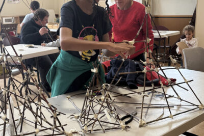 photo d'une jeune fille debout devant une table qui assemble sa lanterne magique faite de branches fines accrochées ensemble par des scotchs - Agrandir l'image 5 sur 8, fenêtre modale