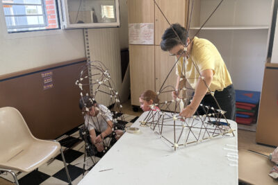 photo d'une animatrice et de deux jeunes filles en train de monter leur lanterne, faite de branches fines accrochées ensemble par des scotchs - Agrandir l'image 2 sur 8, fenêtre modale