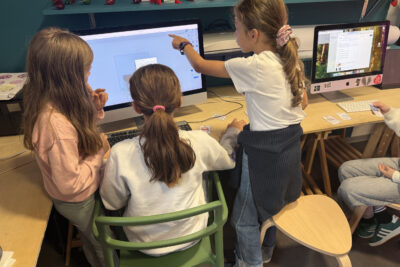 trois jeunes filles travaillent sur un mac, une est assise et contrôle la souris, les deux autres sont debout de chaque côté, celle qui est à sa droite montre quelque chose sur l'écran - Agrandir l'image 5 sur 8, fenêtre modale