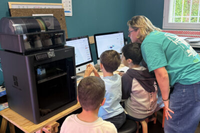 photo de jeunes garçons installés à travailler sur des macs, une jeune femme derrière regarde avec eux ce qu'ils font à l'écran - Agrandir l'image 7 sur 8, fenêtre modale
