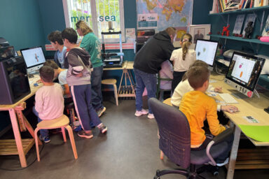 photo de la pièce où sont installés une dizaine de jeunes enfants en train de travailler sur les macs avec l'aide de deux animateurs