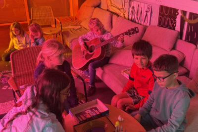 photo portrait dans une pièce éclairée de lumière rose et rouge, avec des canapés, des enfants jouent à un jeu de société pendant qu'un autre joue de la guitare à côté, à l'arrière plan, des jeunes filles jouent à un jeu aussi, par terre - Agrandir l'image 2 sur 8, fenêtre modale