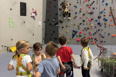 Photo d'un groupe de cinq enfants qui regardent une jeune fille monter au mur d'escalade, ils sont tous équipés d'un baudrier - Agrandir l'image 1 sur 9, fenêtre modale