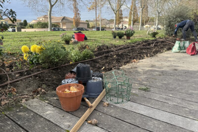 un homme est penché au-dessus d'une zone de terre fraîche le long d'une allée en bois dans un parc, on voit des outils de jardinage et des pots de fleurs vides à côté - Agrandir l'image 2 sur 2, fenêtre modale