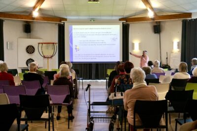 Photo d'une conférence, un diaporama est projeté dans une salle, plusieurs personnes âgées sont assises et écoutent une intervenante - Agrandir l'image 3 sur 20, fenêtre modale