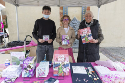 photo d'un stand avec trois personnes derrière, dont deux jeunes, les trois tiennent et montrent des BDs/mangas. Sur la table, d'autres ouvrages (mangas, BDs...) - Agrandir l'image 16 sur 21, fenêtre modale