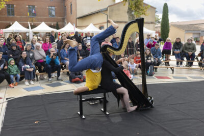 photo du spectacle, la femme joue de la harpe assise et l'homme fait une acrobatie derrière elle, le public assiste au spectacle sur de petits bancs - Agrandir l'image 14 sur 21, fenêtre modale