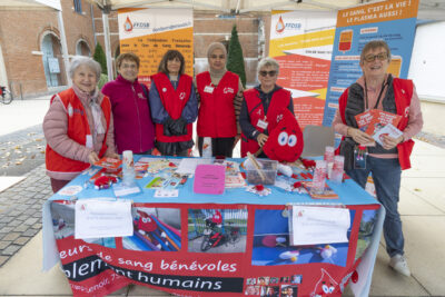 photo du stand de l'association du don du sang, 6 femmes derrière le stand debout qui sourient à l'objectif, vêtues du gilet rouge de leur association, sur la table de la documentation - Agrandir l'image 11 sur 21, fenêtre modale