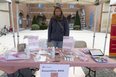 Photo d'un stand associatif pour l'ARSE, une dame derrière la table est debout et sourit à l'objectif, sur la table couverte d'une nappe rose, de la documentation et des produits (crèmes) - Agrandir l'image 4 sur 21, fenêtre modale