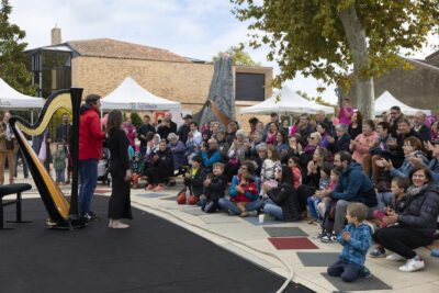 photo d'un spectacle sur le parvis, un homme et une femme sont debout sur une sorte de tapis noir qui fait office de scène, il y a une grande harpe derrière eux. Le public est rassemblé devant, nombreux avec beaucoup d'enfants au premier rang - Agrandir l'image 3 sur 21, fenêtre modale