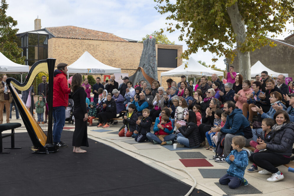 photo d'un spectacle sur le parvis, un homme et une femme sont debout sur une sorte de tapis noir qui fait office de scène, il y a une grande harpe derrière eux. Le public est rassemblé devant, nombreux avec beaucoup d'enfants au premier rang
