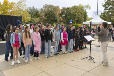 photo d'une chorale de jeunes, alignés en deux rangées sur le parvis, une cheffe de chœur devant eux tandis qu'ils chantent - Agrandir l'image 7 sur 21, fenêtre modale