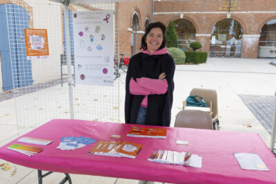 Photo d'un stand, une femme derrière se tient debout, bras croisés et sourit à l'objectif, sur la table on voit de la documentation sur les violences conjugales - Agrandir l'image 5 sur 21, fenêtre modale