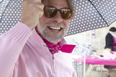 photo d'un homme souriant qui porte des lunettes de soleil et un pull rose clair, avec un ruban rose dessus, il tient un parapluie noir à pois blancs - Agrandir l'image 9 sur 21, fenêtre modale