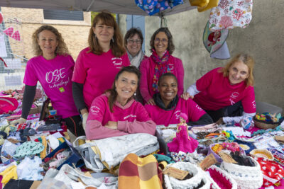 Photo d'un stand, sept femmes habillées en rose derrière la table recouverte de bonnets et autres vêtements et petits objets - Agrandir l'image 2 sur 21, fenêtre modale