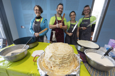 photo de bénévoles en vert avec tablier gris, et d'une assiette pleine de crepes devant eux posée sur une table - Agrandir l'image 64 sur 68, fenêtre modale