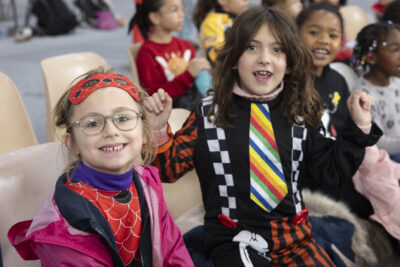 photo de deux jeunes filles déguisées assises dans le public, une est en tenue de rouge avec des écailles, l'autre porte une cravate multicolore et un pantalon à rayures, des bretelles à carreaux blancs et noirs - Agrandir l'image 58 sur 68, fenêtre modale