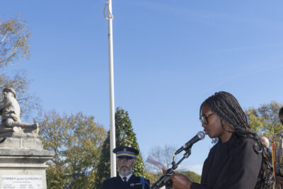 photo d'une jeune qui lit un discours, on voit le monument aux morts et le drapeau français hissé derrière elle - Agrandir l'image 31 sur 38, fenêtre modale