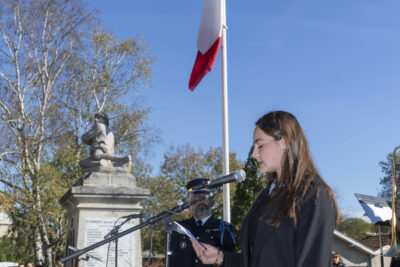 photo d'une jeune qui lit un discours, on voit le monument aux morts et le drapeau français hissé derrière elle - Agrandir l'image 32 sur 38, fenêtre modale