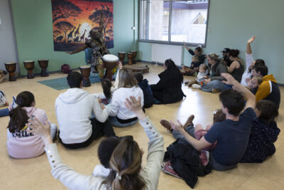 des familles sont rassemblées, assises dans une pièce face à un percussionniste - Agrandir l'image 13 sur 32, fenêtre modale