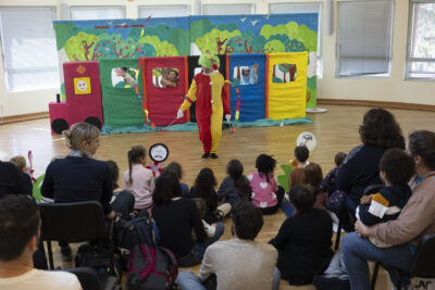 photo d'un spectacle prise de derrière les spectateurs dans une salle, on voit des enfants assis au premier rang, et un décor de train coloré avec des personnes derrière chaque wagon, et un clown entre le train et les spectateurs - Agrandir l'image 2 sur 32, fenêtre modale