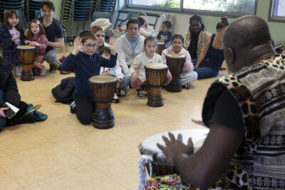 photo des enfants avec leurs parents qui font de la percussion avec des djembés, face à un homme qui en joue lui-même - Agrandir l'image 12 sur 32, fenêtre modale