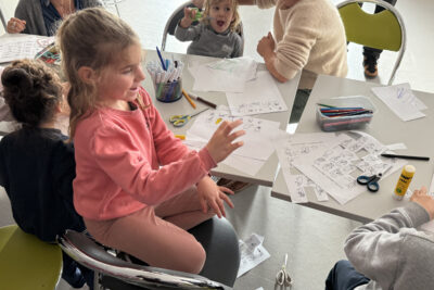 photo d'une petite fille qui fait un signe de main à sa mère, assise sur une chaise, à côté d'elle une table avec des feuilles éparpillées et de l'autre côté un petit enfant bouche grande ouverte - Agrandir l'image 31 sur 32, fenêtre modale