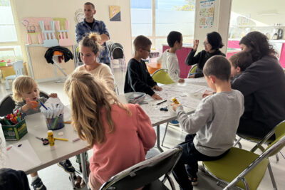 photo d'un atelier avec des enfants et quelques parents réunis dans une salle autour d'une table, faisant des signes avec la main ou découpant des feuilles - Agrandir l'image 30 sur 32, fenêtre modale
