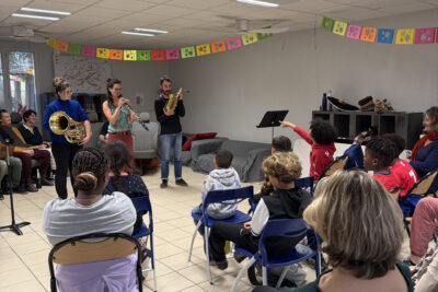 photo d'un petit concert dans une salle d'école, avec des enfants assis qui regardent des musiciens, un enfant a le doigt tendu vers un instrument - Agrandir l'image 26 sur 32, fenêtre modale