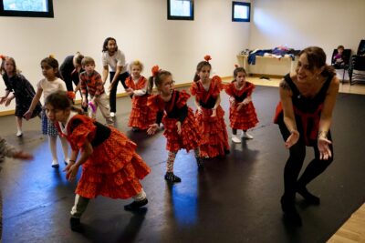 photo du groupe d'enfants, une dizaine, qui s'entraînent à danser en imitant l'adulte devant, en faisant un geste des mains ouvertes, le corps penché sur le côté - Agrandir l'image 6 sur 7, fenêtre modale