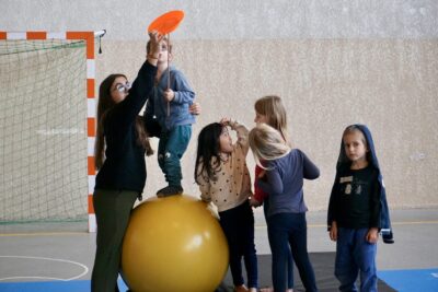 une jeune femme aide un jeune garçon à tenir en équilibre debout sur un gros ballon orange tout en tenant un baton en bois surmonté d'une assiette orange - Agrandir l'image 14 sur 16, fenêtre modale