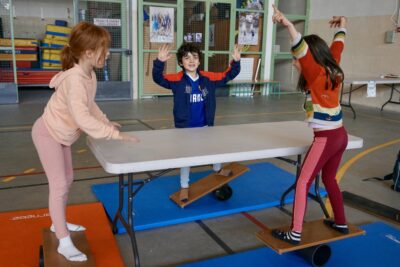 deux jeunes filles et un jeune garçon tiennent en équilibre sur une planche posée sur un rondin, deux ont les mains en l'air et ne s'aident pas de la table devant eux pour garder l'équilibre - Agrandir l'image 13 sur 16, fenêtre modale